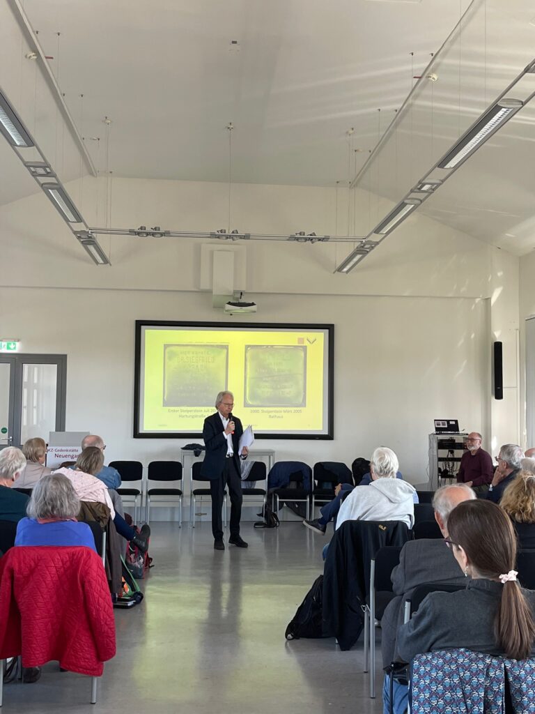 Ingo Wille spricht bei der Mitgliederversammlung. Auf der Leinwand hinter ihm sind Fotos der ersten und der 1000. Stolpersteinverlegung in Hamburg zu sehen.