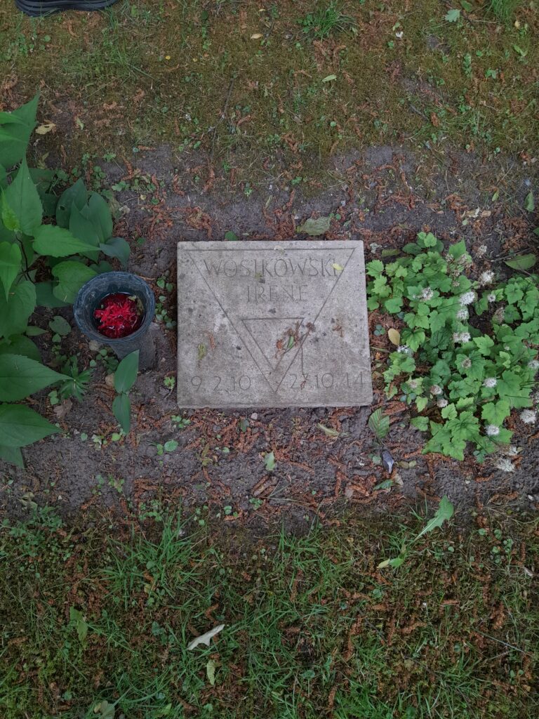 Flacher Gedenkstein mit Winkel und der Inschrift „Wosikowski Irene“, daneben ein Blumenbehälter, eingebettet in Erde und Gras.