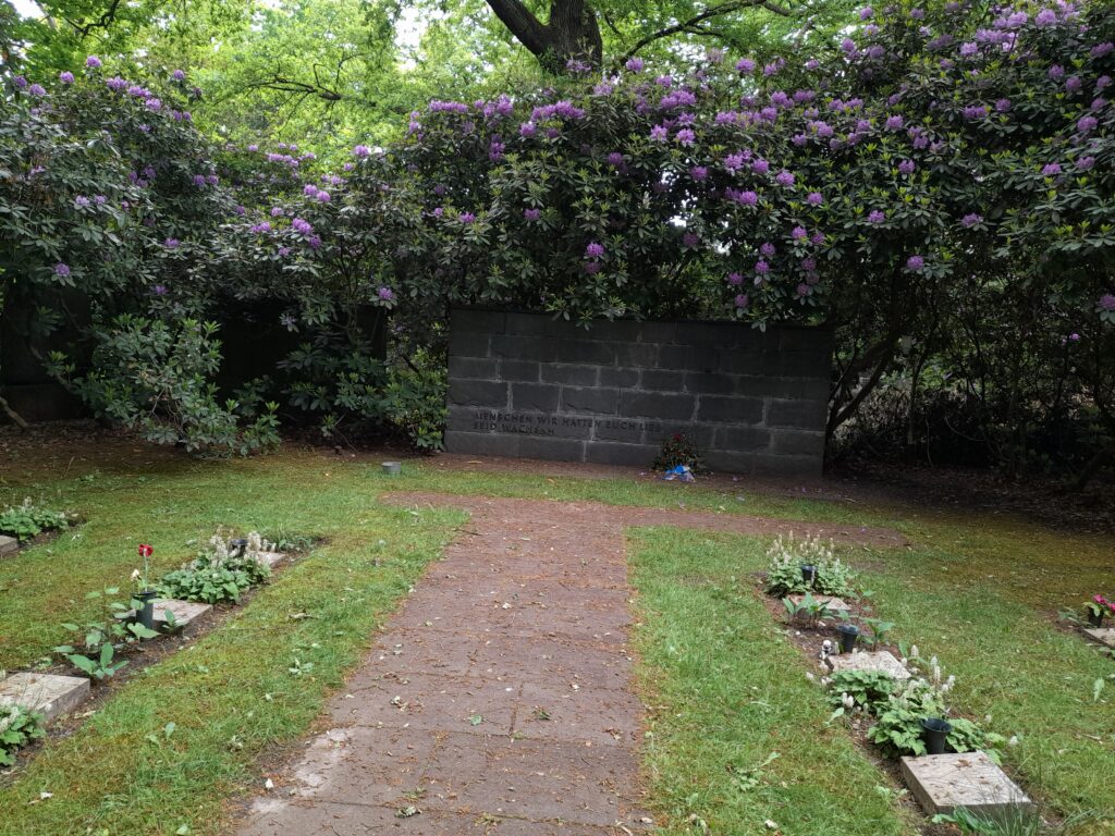 Steinmauer mit eingemeißelter Inschrift „Menschen wir hatten euch lieb / seid wachsam“, umrahmt von blühenden Rhododendren. Davor gepflegte Grabplatten und ein Weg
