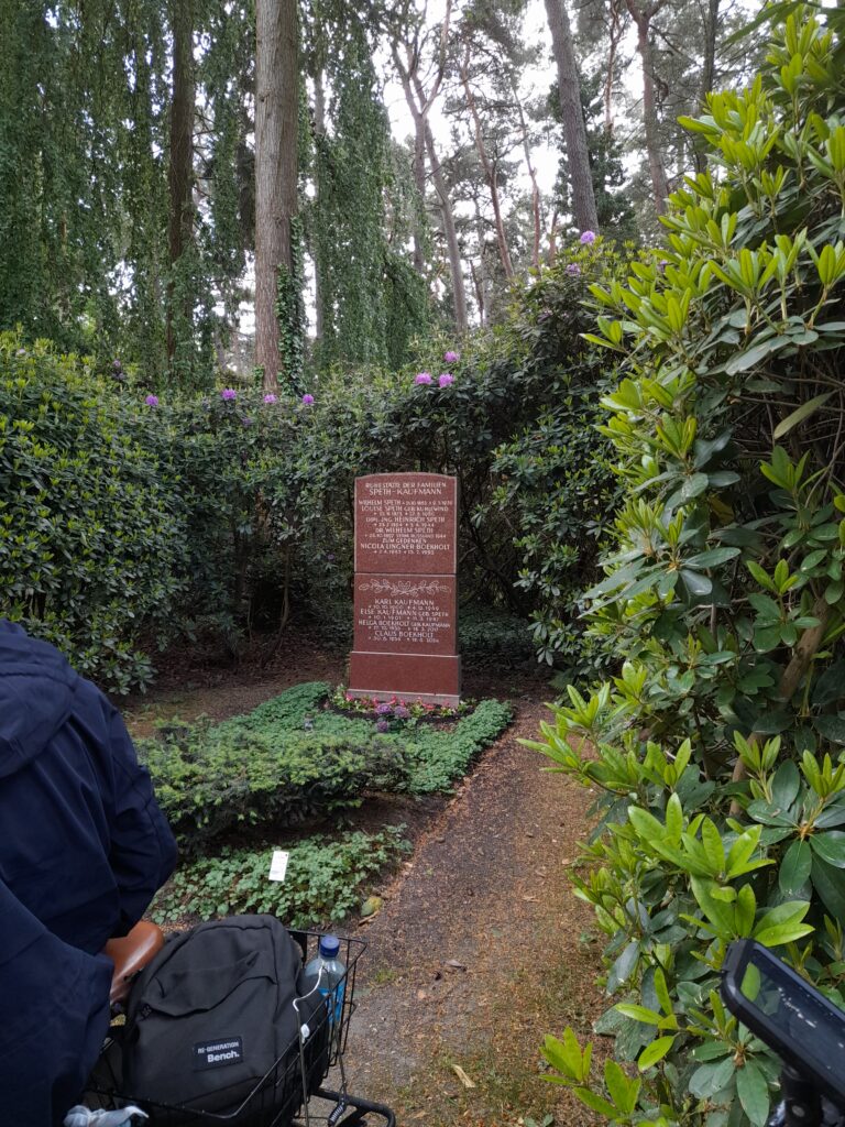 Roter Grabstein mit Inschrift für Mitglieder der Familie Speth-Kaufmann, darunter auch Karl Kaufmann, eingebettet in grüne Pflanzen und blühende Rhododendren, umgeben von Bäumen.