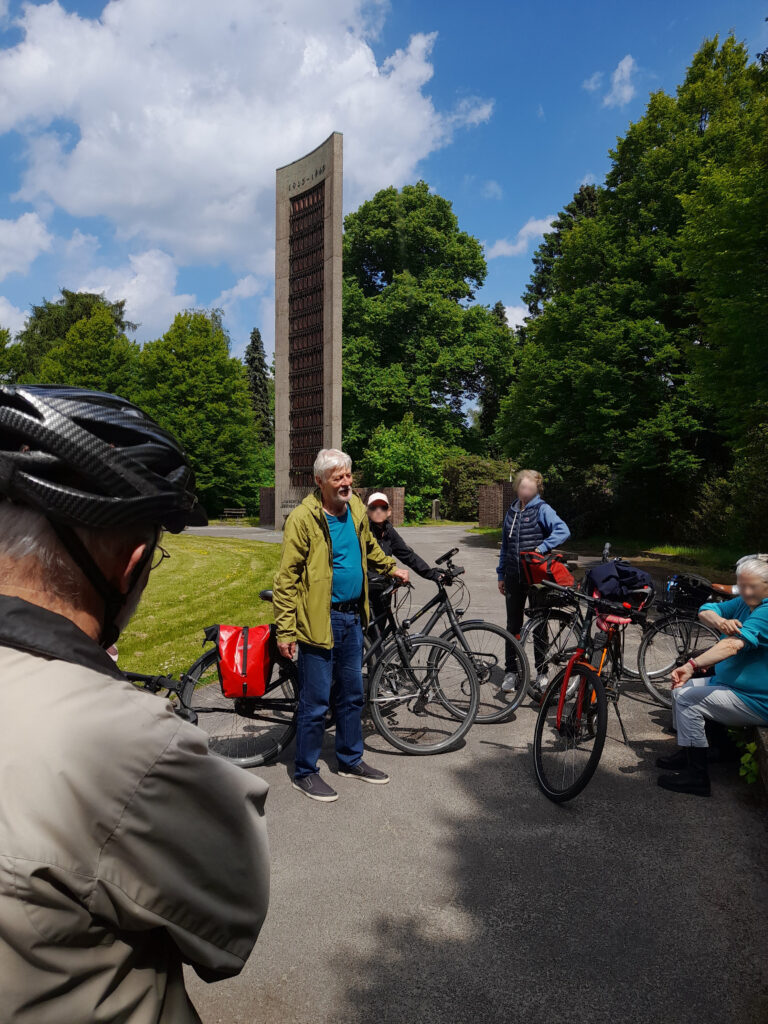 Menschen mit Fahrrädern versammeln sich vor einem hohen Mahnmal. Eine Person spricht, andere stehen oder sitzen im Halbkreis und hören zu. Bäume und blauer Himmel im Hintergrund.