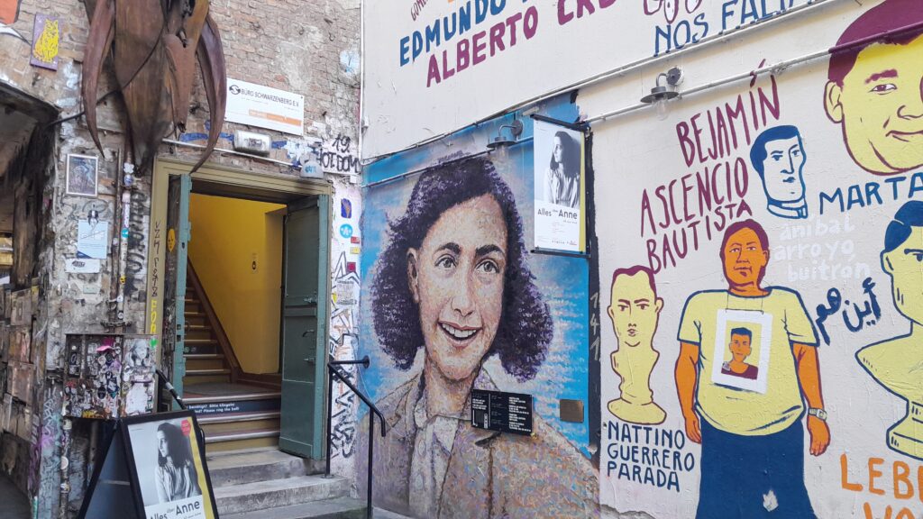 Eingang zum Anne Frank Zentrum in Berlin mit Treppenhaus hinter einer offenen Tür und einem großen, farbigen Porträt von Anne Frank als Wandgemälde neben dem Eingang.