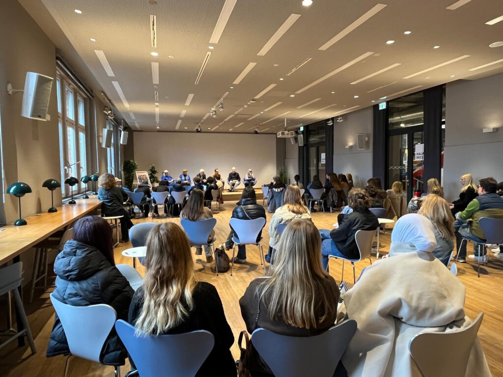 Gefüllter Saal mit Jugendlichen in der Rückansicht auf blauen Stühlen, im Hintergrund Bühne mit sechs Personen