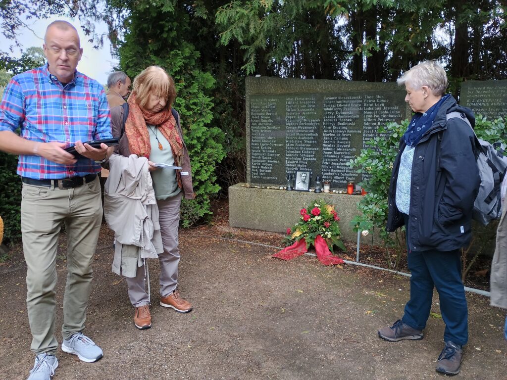Teilnehmend der Gedenkstättenfahrt vor der Namenstafel der im KZ Engerhafe umgekommenen Häftlinge auf dem Friedhof des Ortes, Foto: Barbara Hartje, Engerhafe, 2026