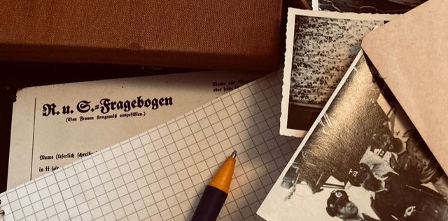 Historische Dokumente und Fotos liegen übereinander, in der Mitte ein kariertes Blatt Papier mit einem Kugelschreiber